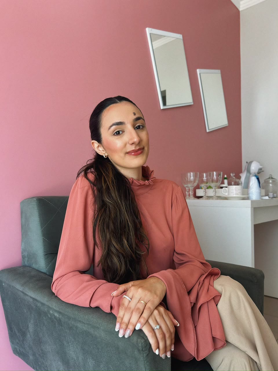 Tainá Quevedo sorrindo, sentada na poltrona cinza do consultório de parede rosa